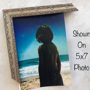May include: A silver-colored ornate picture frame partially displaying a 5x7 inch photo. The photo shows a person standing on a beach, facing the ocean. The sky is blue, and the ocean is visible in the background.