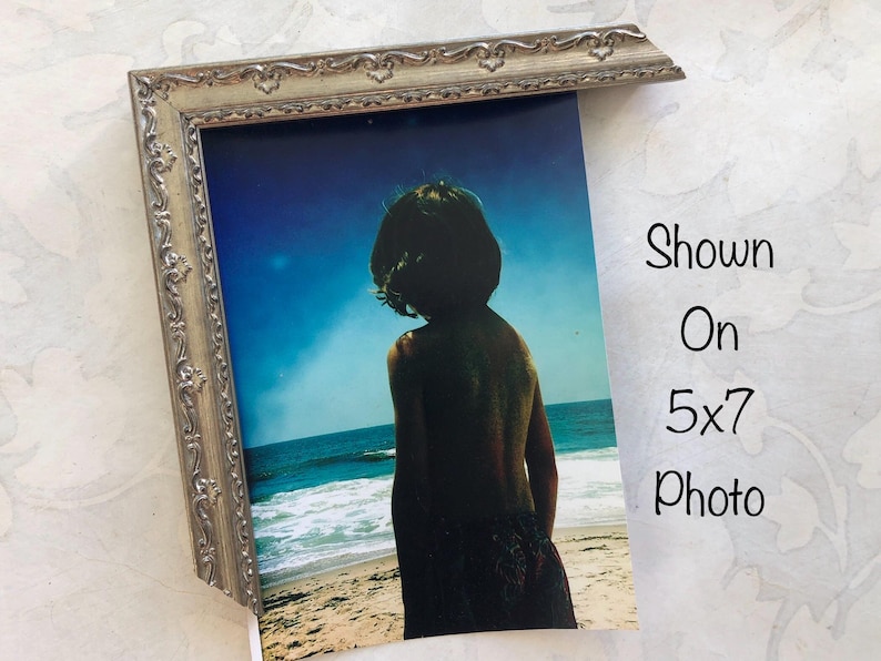 May include: A silver-colored ornate picture frame partially displaying a 5x7 inch photo. The photo shows a person standing on a beach, facing the ocean. The sky is blue, and the ocean is visible in the background.