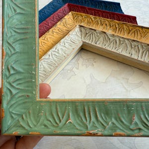 May include: A collection of decorative picture frames in various colors, including green, yellow, red, and blue. The frames have a carved leaf pattern and a distressed finish, adding a vintage aesthetic. The frames are made of wood.