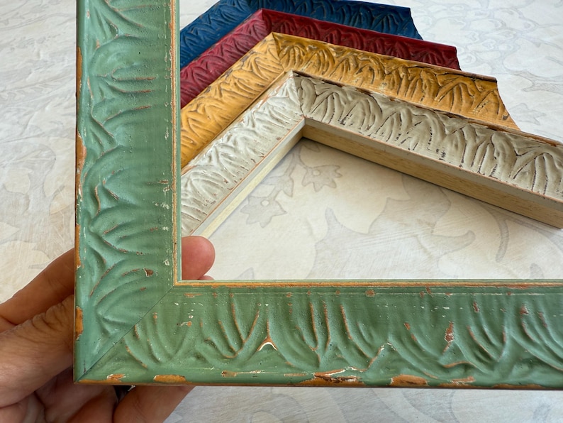 May include: A collection of decorative picture frames in various colors, including green, yellow, red, and blue. The frames have a carved leaf pattern and a distressed finish, adding a vintage aesthetic. The frames are made of wood.