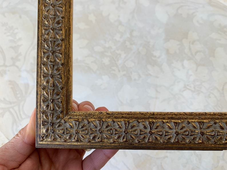May include: Close up of a corner of a gold picture frame with an ornate design. The frame has a raised edge and a detailed pattern of leaves and flowers.