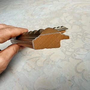 May include: A cross-section of a brown wooden picture frame with a textured, light brown interior. The frame has a decorative, ridged edge and a metallic accent. The frame is held in a hand.