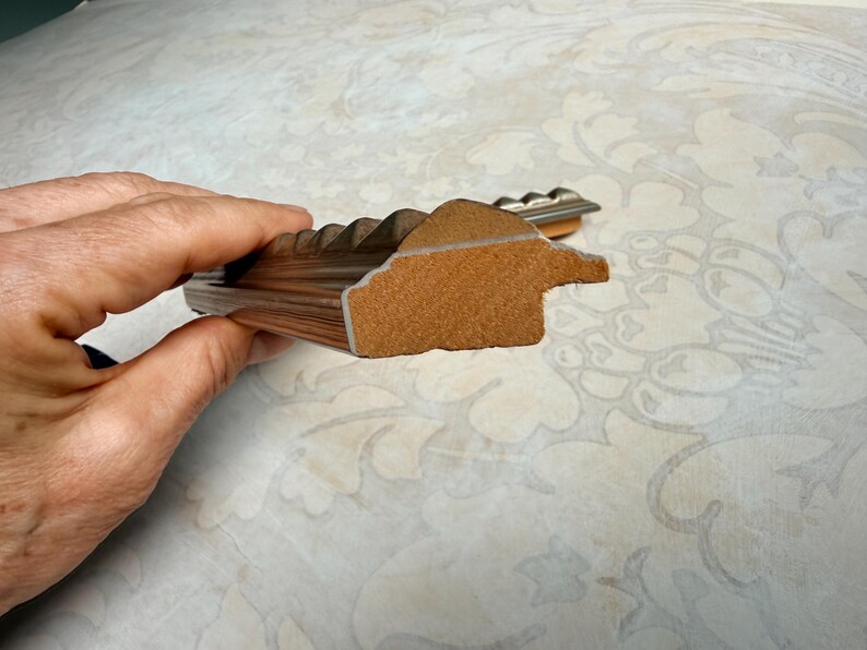 May include: A cross-section of a brown wooden picture frame with a textured, light brown interior. The frame has a decorative, ridged edge and a metallic accent. The frame is held in a hand.