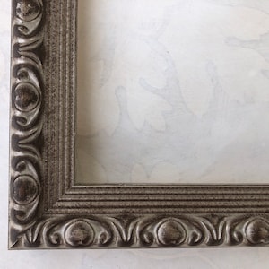 May include: A silver picture frame with an ornate design. The frame has a raised edge and a decorative border.