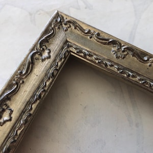 May include: Close-up of a corner of a decorative picture frame. The frame is a muted gold color with ornate, raised scrollwork and floral details. The frame's design is vintage and elegant.