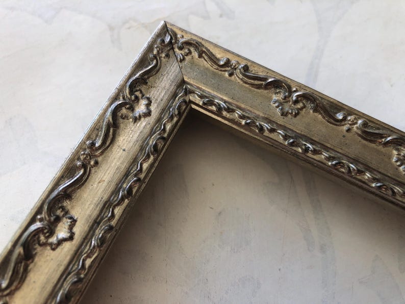May include: Close-up of a corner of a decorative picture frame. The frame is a muted gold color with ornate, raised scrollwork and floral details. The frame's design is vintage and elegant.