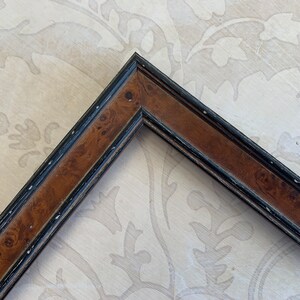 May include: A corner of a picture frame with a brown wood finish and a black painted border. The wood has a burl pattern.