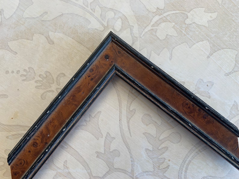 May include: A corner of a picture frame with a brown wood finish and a black painted border. The wood has a burl pattern.