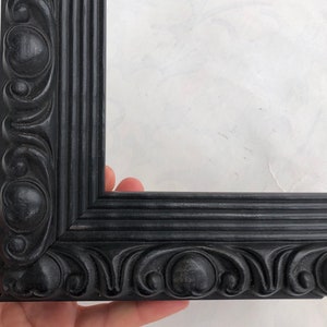 K&ouml;nnte beinhalten: Schwarzer, verzierter Bilderrahmen mit einem detaillierten, geschnitzten Design. Der Rahmen hat eine glatte, schwarze Oberfl&auml;che.