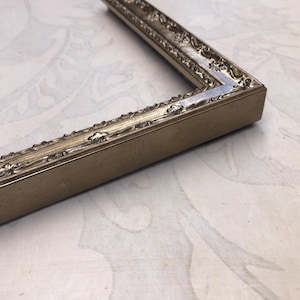 May include: Close-up of a section of a rectangular picture frame. The frame is a muted gold color with ornate detailing along the inner and outer edges. The corner of the frame is visible, showing the construction and the decorative elements.
