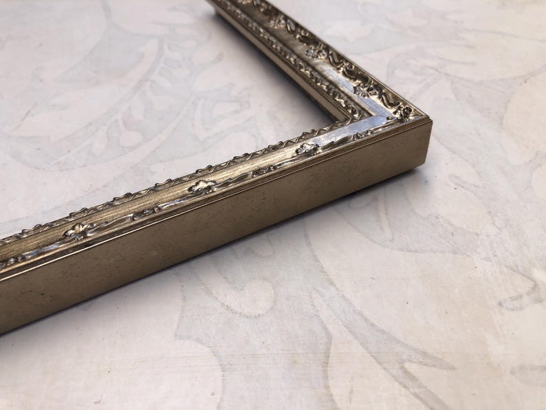 May include: Close-up of a section of a rectangular picture frame. The frame is a muted gold color with ornate detailing along the inner and outer edges. The corner of the frame is visible, showing the construction and the decorative elements.