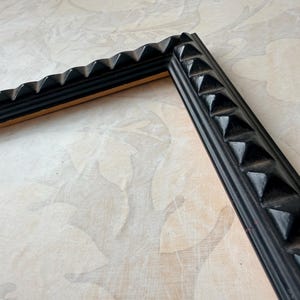 May include: Two pieces of a black picture frame with a textured, geometric design. The frame has a series of triangular shapes along the edges, creating a decorative border. The frame is set against a light-colored background.