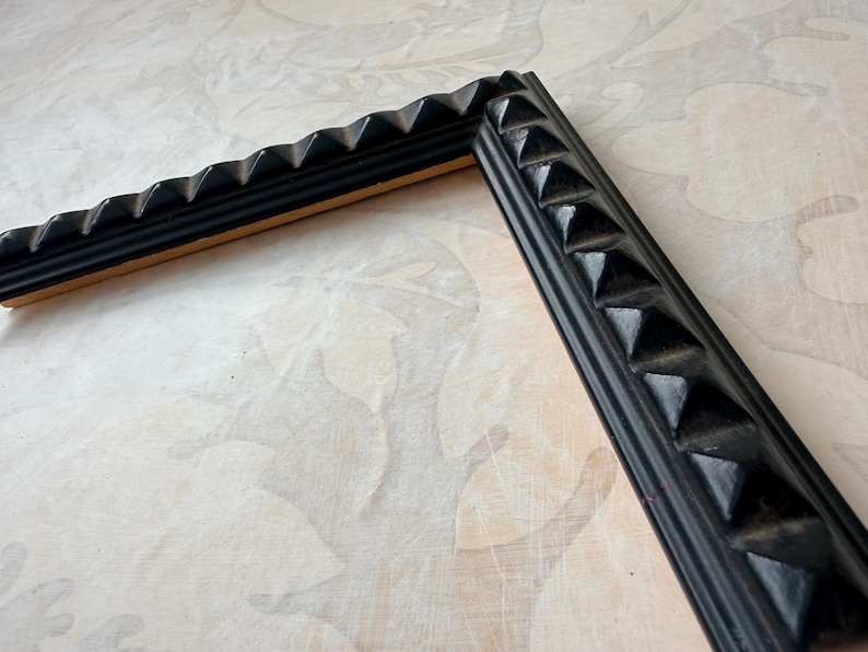 May include: Two pieces of a black picture frame with a textured, geometric design. The frame has a series of triangular shapes along the edges, creating a decorative border. The frame is set against a light-colored background.