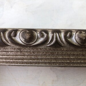 May include: Close-up of a silver picture frame with a decorative, ornate border. The frame has a distressed, aged look with a dark gray patina.