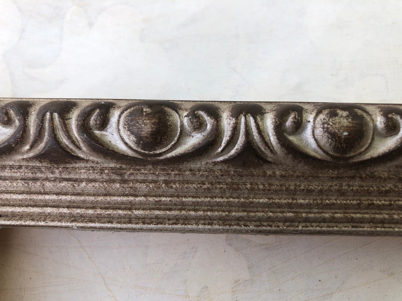May include: Close-up of a silver picture frame with a decorative, ornate border. The frame has a distressed, aged look with a dark gray patina.