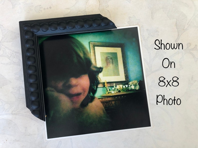 May include: A black picture frame with a beaded border surrounds a photograph of a young person with dark hair. The photo shows the person looking down and smiling. The photo is displayed on an 8x8 photo.