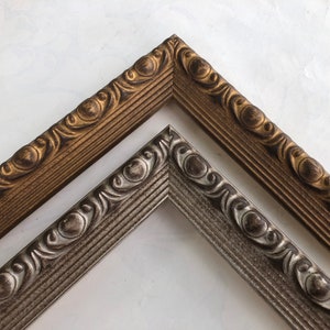May include: Two ornate picture frames, one gold and one silver, with a decorative, raised design. The frames are angled to show the detail of the design.