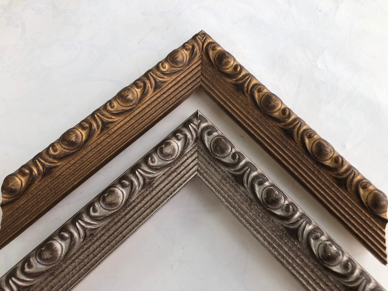 May include: Two ornate picture frames, one gold and one silver, with a decorative, raised design. The frames are angled to show the detail of the design.