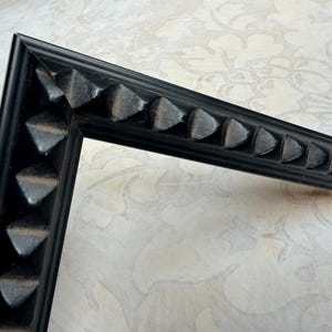 May include: A corner of a black picture frame with a decorative, repeating triangular pattern along the edges. The frame is made of a dark material and is set against a patterned, off-white background.