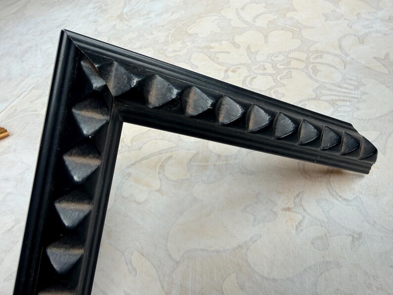 May include: A corner of a black picture frame with a decorative, repeating triangular pattern along the edges. The frame is made of a dark material and is set against a patterned, off-white background.