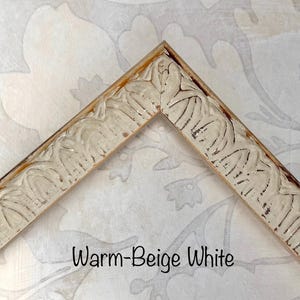 May include: A corner of a warm-beige white picture frame. The frame has a carved, ornate design with a distressed finish. The text "Warm-Beige White" is displayed in a black font.