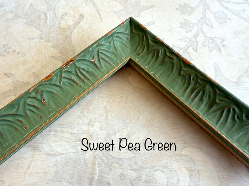 May include: A corner of a Sweet Pea Green picture frame. The frame has a distressed finish with carved leaf details. The frame is a muted green color with hints of brown. The text "Sweet Pea Green" is written in a script font.