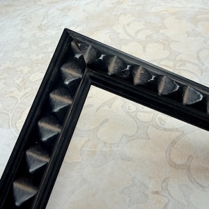 May include: A black picture frame corner with a decorative, textured edge. The frame's edge features a series of raised, triangular shapes. The frame is made of a dark material and is set against a patterned background.