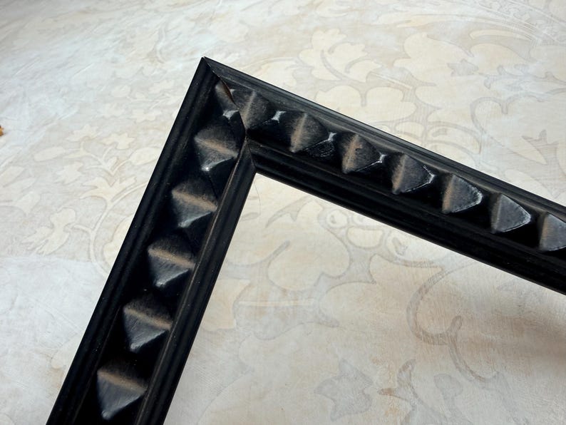 May include: A black picture frame corner with a decorative, textured edge. The frame's edge features a series of raised, triangular shapes. The frame is made of a dark material and is set against a patterned background.