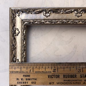 May include: Ornate silver-toned picture frame with decorative floral and scroll detailing. The frame is rectangular and includes a wooden ruler with text that reads "R. E. Smith Cherry 1912".