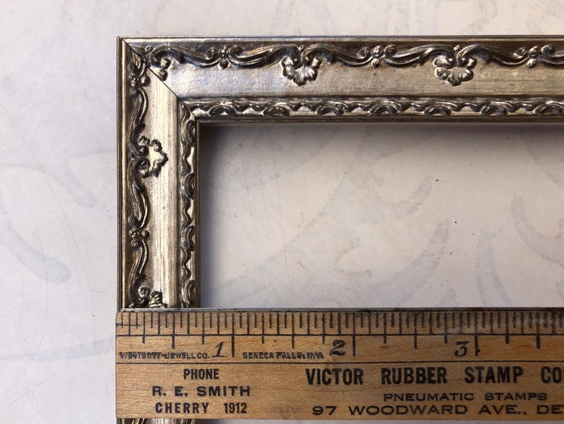 May include: Ornate silver-toned picture frame with decorative floral and scroll detailing. The frame is rectangular and includes a wooden ruler with text that reads "R. E. Smith Cherry 1912".