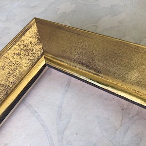 May include: Close-up of a corner of a gold picture frame. The frame has a textured, distressed finish with dark brown accents. The frame's interior is black, and the background is a light, patterned surface.