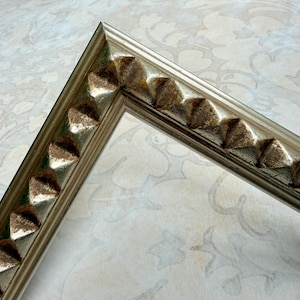 May include: Silver-toned picture frame corner with decorative, textured accents. The frame features a series of raised, pyramid-shaped embellishments along the edge, adding a touch of elegance. The frame's design suggests a vintage or antique style, suitable for displaying photographs or artwork.