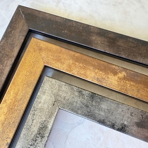 May include: Close-up of three picture frames with textured surfaces. The frames are in shades of bronze, gold, and silver, with a distressed finish. The frames are angled, showcasing their corner details and layered design.