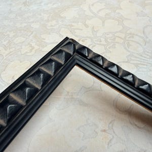 May include: A corner of a black picture frame with a textured, geometric design. The frame has a series of raised, pyramid-shaped elements along its outer edge, creating a decorative border. The frame is set against a patterned background.