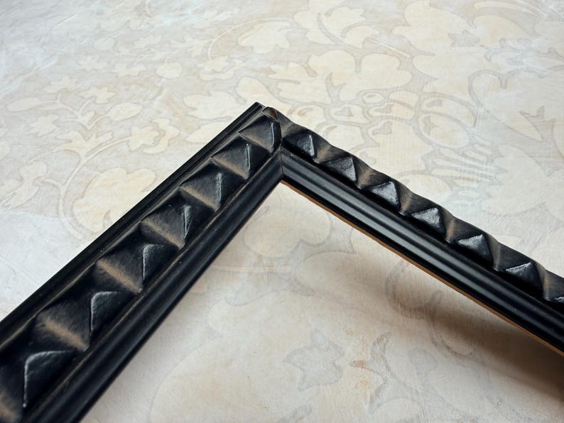 May include: A corner of a black picture frame with a textured, geometric design. The frame has a series of raised, pyramid-shaped elements along its outer edge, creating a decorative border. The frame is set against a patterned background.