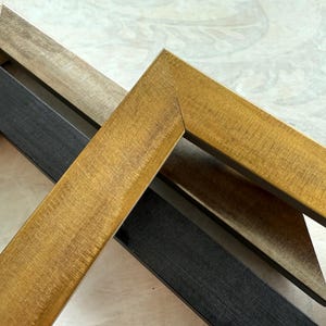 May include: Close-up of three wooden picture frames in varying colors. One frame is a light tan, another is black, and the third is a warm gold. The frames are angled, showcasing their corners and wood grain.