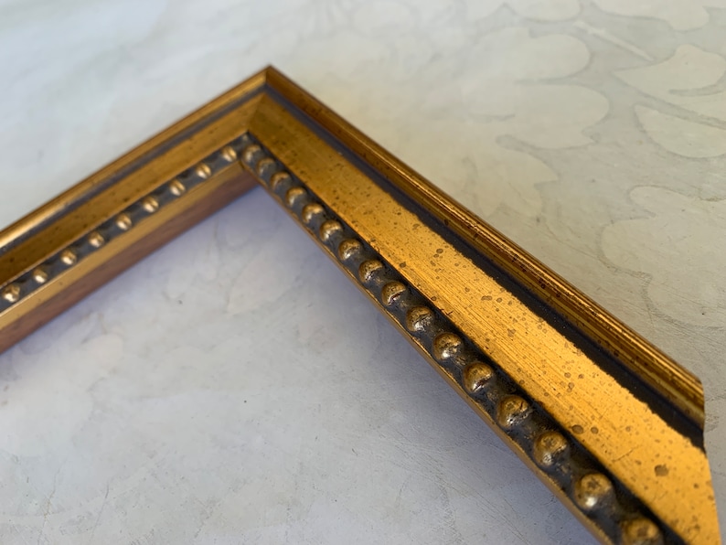 May include: Close-up of a gold picture frame with a beaded edge. The frame is made of wood and has a distressed finish.