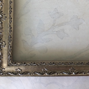 May include: Ornate, gold-toned picture frame with detailed floral and scrollwork. The frame surrounds a light-colored background with a subtle floral pattern. The frame's design suggests a vintage or antique style.