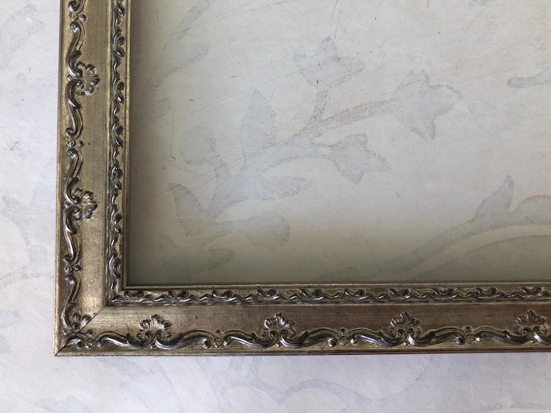 May include: Ornate, gold-toned picture frame with detailed floral and scrollwork. The frame surrounds a light-colored background with a subtle floral pattern. The frame's design suggests a vintage or antique style.