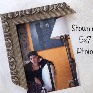 May include: A silver-colored ornate picture frame with a 5x7 photo inside. The photo shows a young woman sitting on a bed with a lamp in the background. The text "Shown on 5x7 Photo" is written in black on a white background.