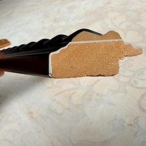 May include: Close-up of a black picture frame with a decorative, ridged edge. The frame's side reveals a light brown core and a white inner edge. The frame is held by a hand, with a patterned surface in the background.