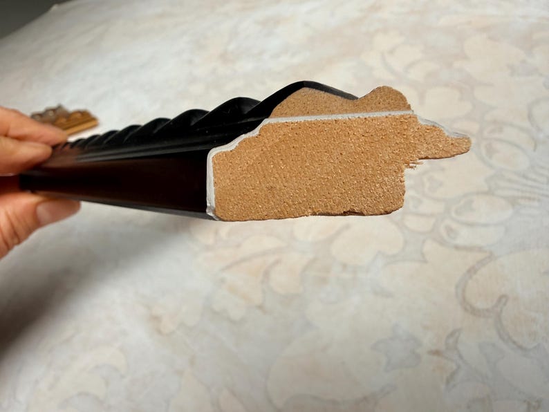 May include: Close-up of a black picture frame with a decorative, ridged edge. The frame's side reveals a light brown core and a white inner edge. The frame is held by a hand, with a patterned surface in the background.