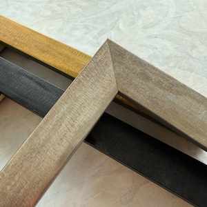 May include: Close-up of three wooden picture frames in gold, black, and gray. The frames are angled, showcasing their textured surfaces and corner joints. The neutral tones suggest a versatile design for various artwork.