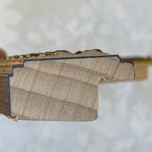 May include: Close-up of the edge of a wooden picture frame with a gold-painted finish. The frame has a decorative, ornate design.