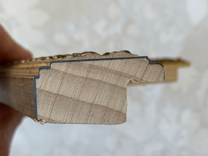 May include: Close-up of the edge of a wooden picture frame with a gold-painted finish. The frame has a decorative, ornate design.