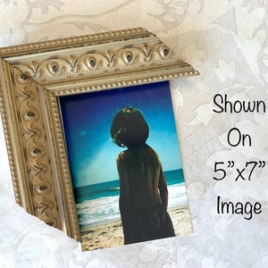 Pu&ograve; includere: Una cornice per foto ornata in tonalit&agrave; dorata, con un'immagine di 5" x 7" di una persona in piedi su una spiaggia. Il testo "Shown On 5" x 7" Image" &egrave; visibile nell'immagine.