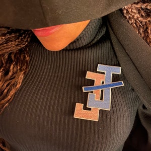 May include: A black mock neck shirt with a pink and blue embroidered patch featuring the letter "J" in a stylized font.