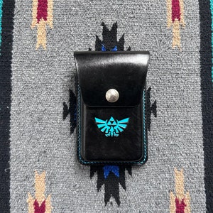 May include: A black leather wallet with a blue Triforce logo. The wallet has a silver button closure and a stitched edge.