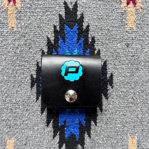 Puede incluir: Una cartera de cuero negro con un logotipo "P" turquesa en forma de nube, asegurada con un broche plateado. La cartera est&aacute; sobre un fondo textil con estampado gris, azul y turquesa.