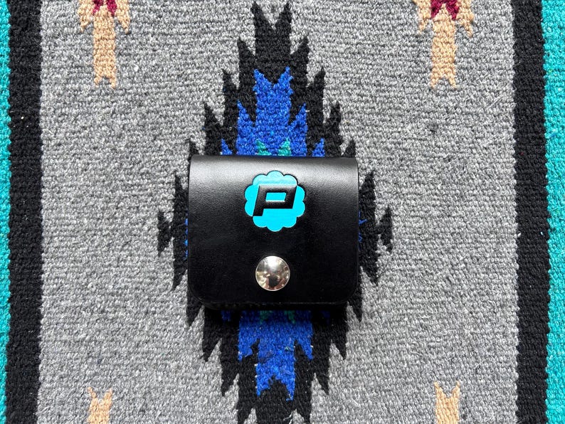 Puede incluir: Una cartera de cuero negro con un logotipo "P" turquesa en forma de nube, asegurada con un broche plateado. La cartera est&aacute; sobre un fondo textil con estampado gris, azul y turquesa.
